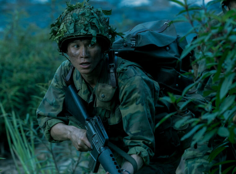Hoa Xuande wearing military camo holding a rifle in the jungle in The Sympathizer | Agents of Fandom