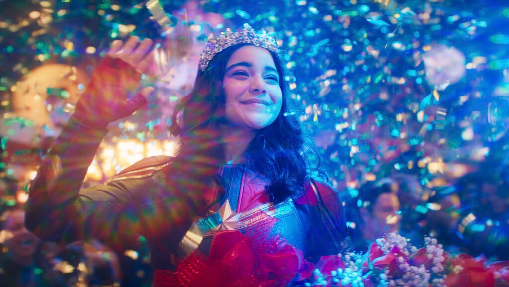Kamala Khan surrounded by glitter and confetti, waving to the crowd with a crown on her head.