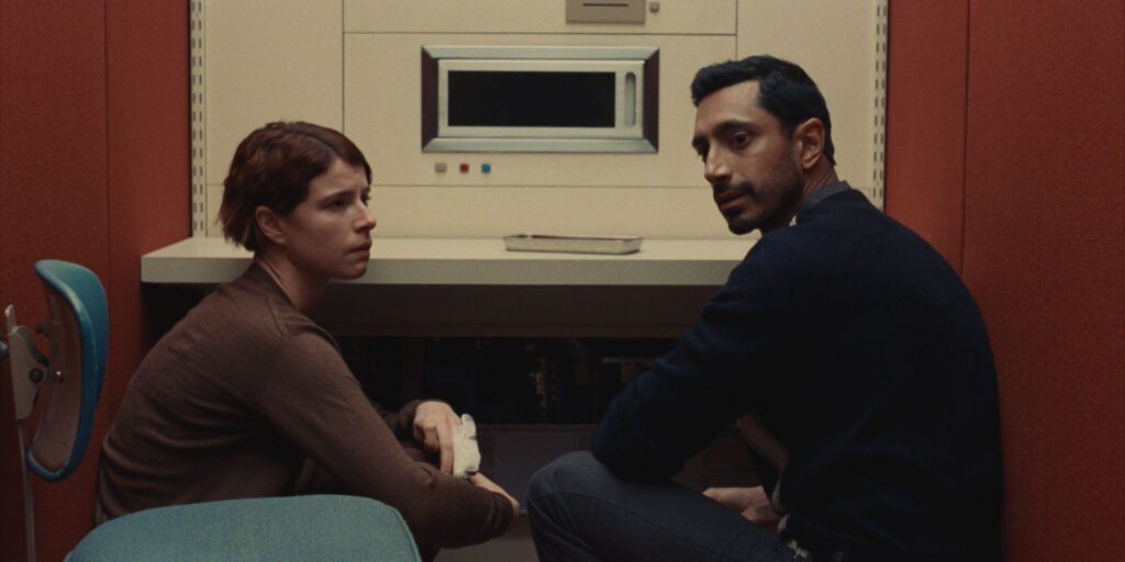 Jessie Buckley and Riz Ahmed in Fingernails are crouching on the ground in front of a beige desk, with red walls behind them | TIFF 2023 | Agents of Fandom