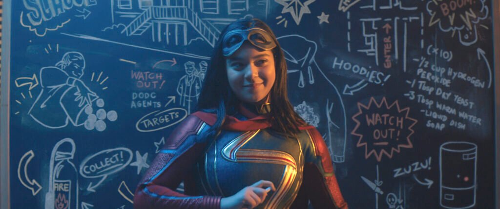 Iman Vellani dressed in her Ms. Marvel hero suit standing behind a blackboard with drawings | Spider-Man team-ups | Agents of Fandom