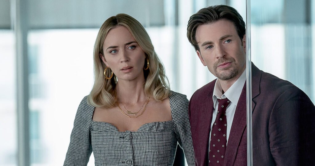 Emily Blunt wearing a salt and pepper colored dress and Chris Evans wearing a maroon suit in a scene in Pain Hustlers | Agents of Fandom