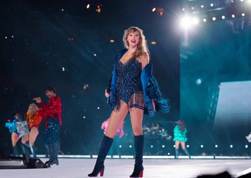 Taylor Swift posing and smiling to the crowd onstage at the Eras Tour | Taylor Swift: The Eras Tour | Agents of Fandom