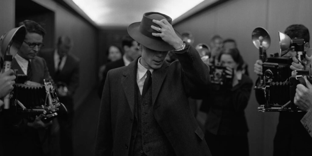 Cillian Murphy as J. Robert Oppenheimer, holding his hat and walking through a room of press | Agents of Fandom