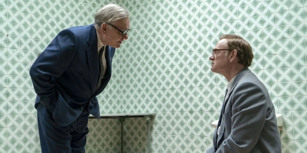 Alan Williams standing up and Jared Harris sitting down in a room together in Chernobyl | Agents of Fandom