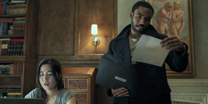 Donald Glover holding a sheet of paper and Maya Erskine sitting at a laptop in Mr. and Mrs. Smith | Agents of Fandom