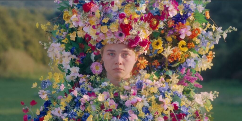 A young woman (Florence Pugh) wears a scared and sad expression on her face. She is covered in flowers of all colors and is surrounded by open green fields. 