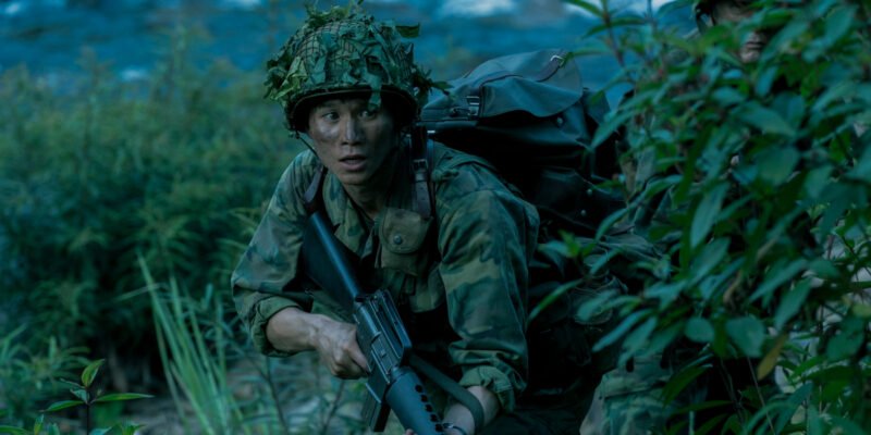 Hoa Xuande wearing military camo holding a rifle in the jungle in The Sympathizer | Agents of Fandom