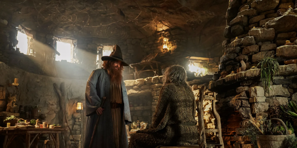 Tom Bombadil standing in front of The Stranger in a cottage in Rings of Power Season 2 | Agents of Fandom