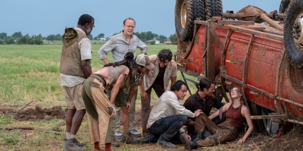 The main cast of Twisters huddles around Daisy Edgar Jones who sits against an overturned truck in a field in Twisters | Agents of Fandom