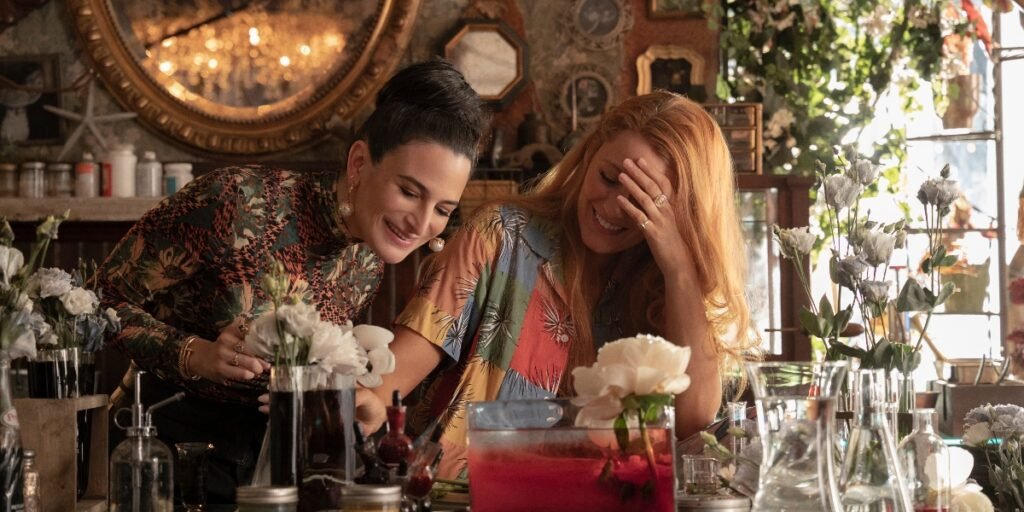 Jenny Slate and Blake Lively wearing warm vibrant clothing inside a floral shop surrounded by flowers and glass containers. They're both looking down at an experiment Lively is doing. | Agents of Fandom