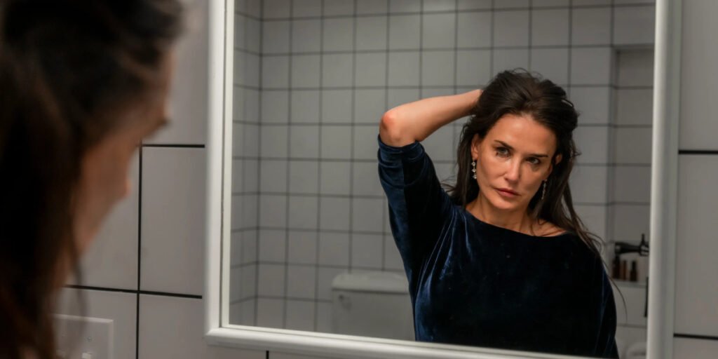 Demi Moore holding her hair while she looks at herself in the mirror in The Substance | Agents of Fandom