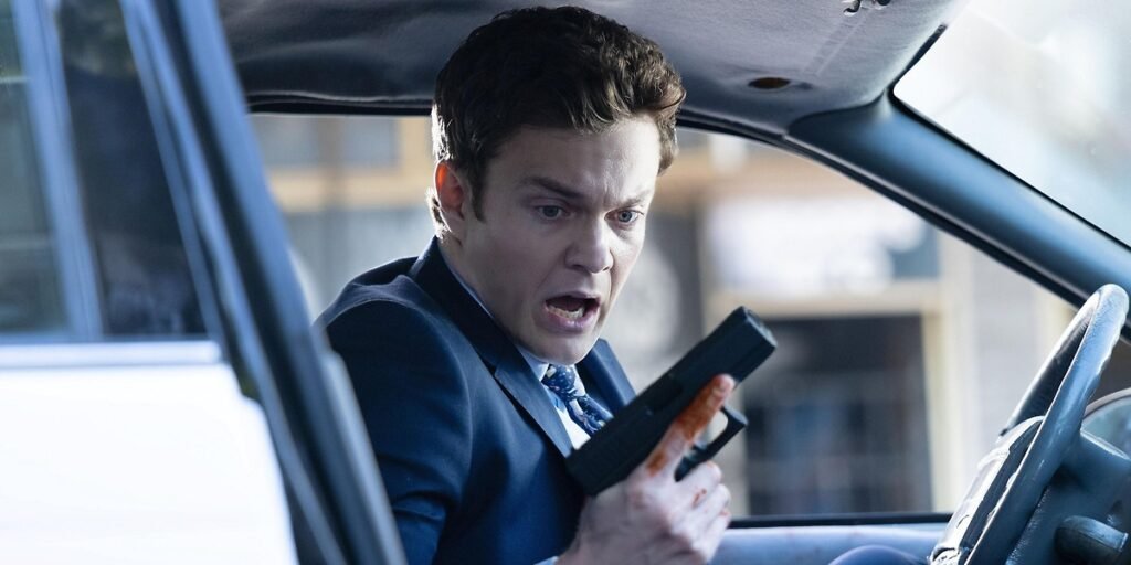 Jack Quaid holding a gun in a car with a gasp on his face I Agents of Fandom