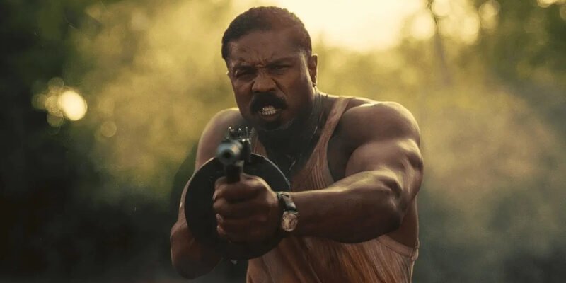 Michael B. Jordan holding a tommy gun with and shouting as he shoots in Sinners | Agents of Fandom